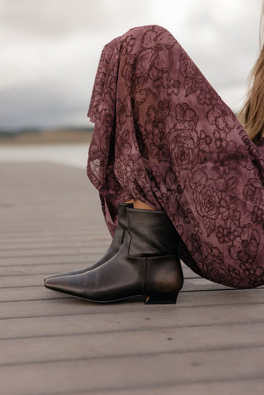 Person wearing black boots and a patterned skirt sitting on a wooden deck.