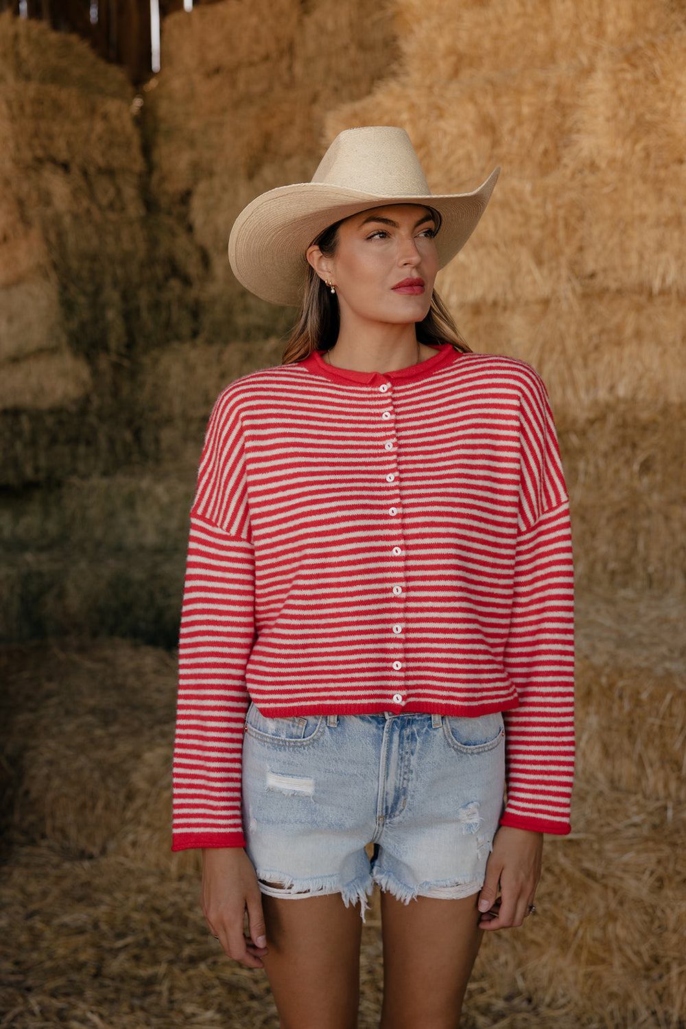 Woman wearing a red and white striped shirt and cowboy hat standing in front of hay bales.