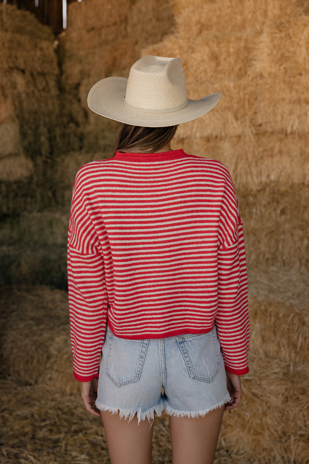 Person wearing a red and white striped shirt and a cowboy hat, standing in front of hay bales.