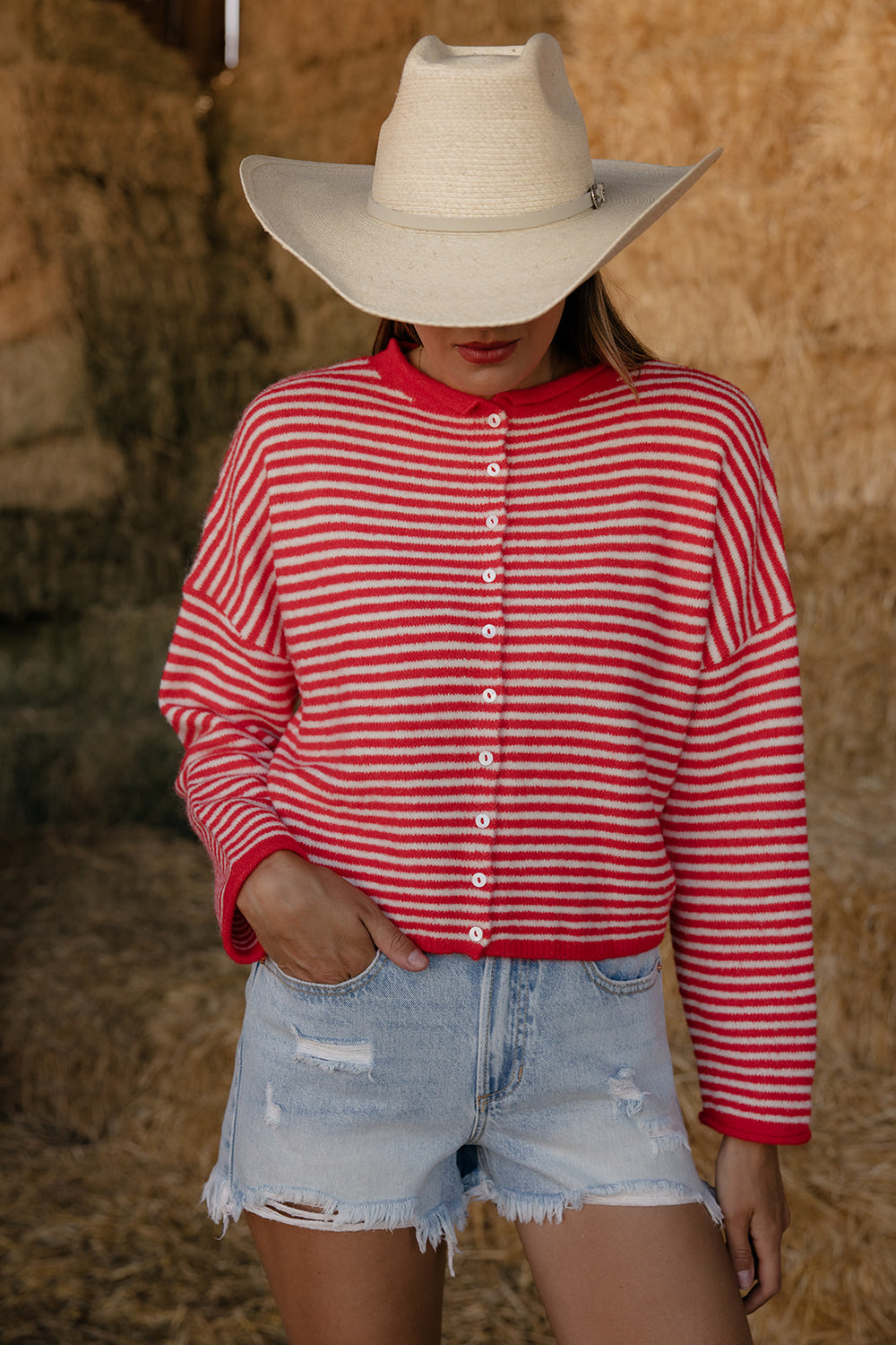 Person wearing a red and white striped shirt, denim shorts, and a cowboy hat in front of hay bales.
