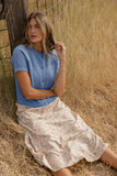Woman in a blue top and patterned skirt sitting in a field of dry grass.