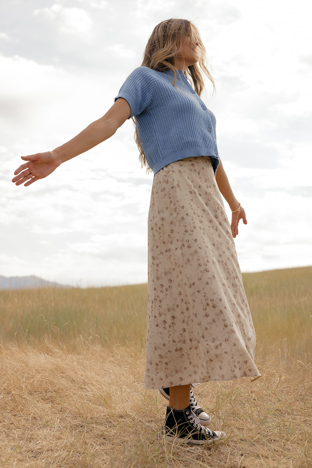 Woman in a blue top and floral skirt standing in a field with outstretched arms.