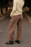 Person wearing a beige sweater and brown pants standing on a road with trees in the background