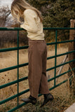 Person wearing a cream sweater and brown pants standing next to a green metal gate with a natural background.