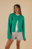 A woman wearing a green button-up cardigan with a rolled collar and arm cuffs, paired with light gray shorts.