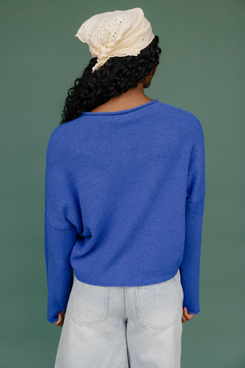 Person wearing a blue sweater and light-colored pants with a headscarf on a green background