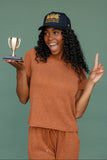 Woman in brown outfit holding a small trophy and pointing upwards against a green background