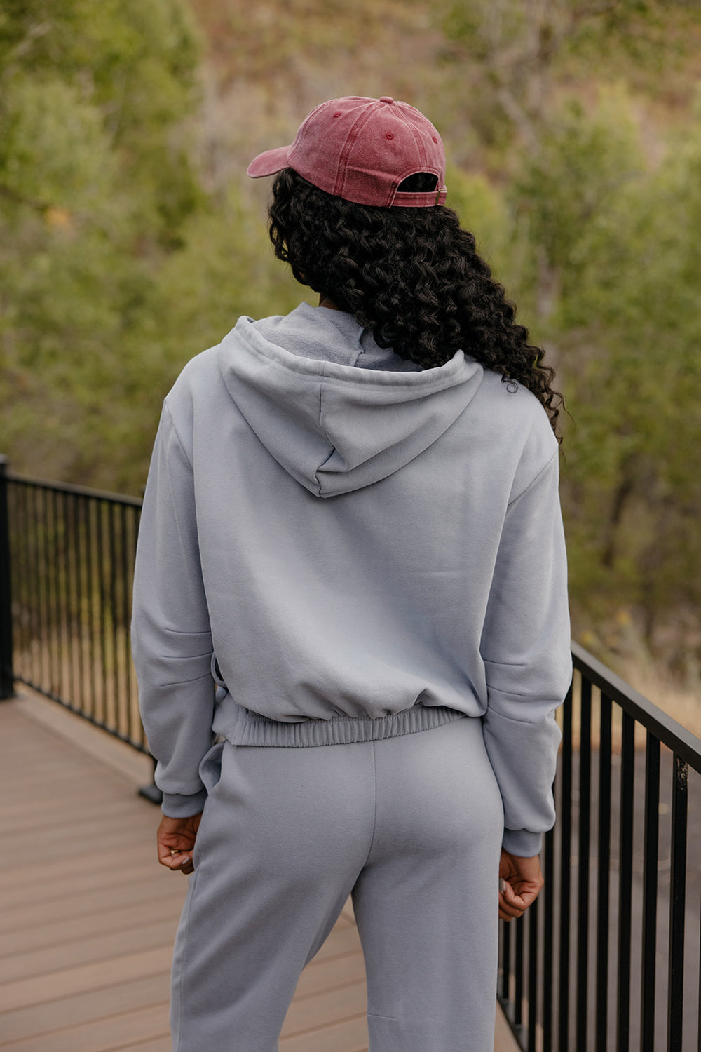 Person wearing a light gray hoodie and pants with a pink cap, standing on a wooden deck with greenery in the background.