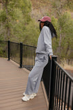 Person wearing a gray tracksuit and pink cap standing on a wooden deck with a forest background