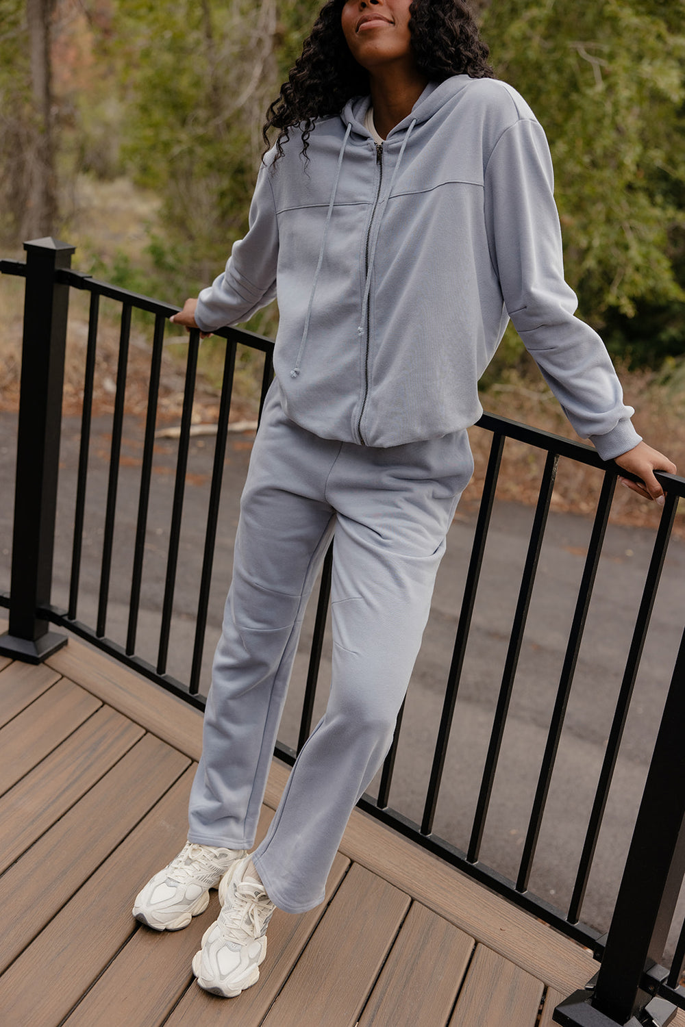 Person wearing a gray tracksuit standing on a wooden deck with greenery in the background