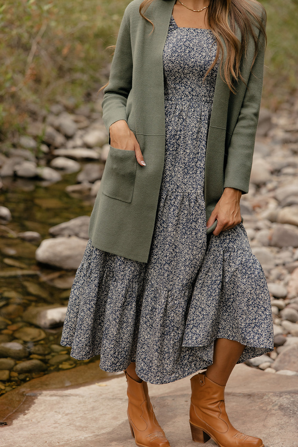 Woman wearing a floral dress with a green cardigan and brown boots by a stream.