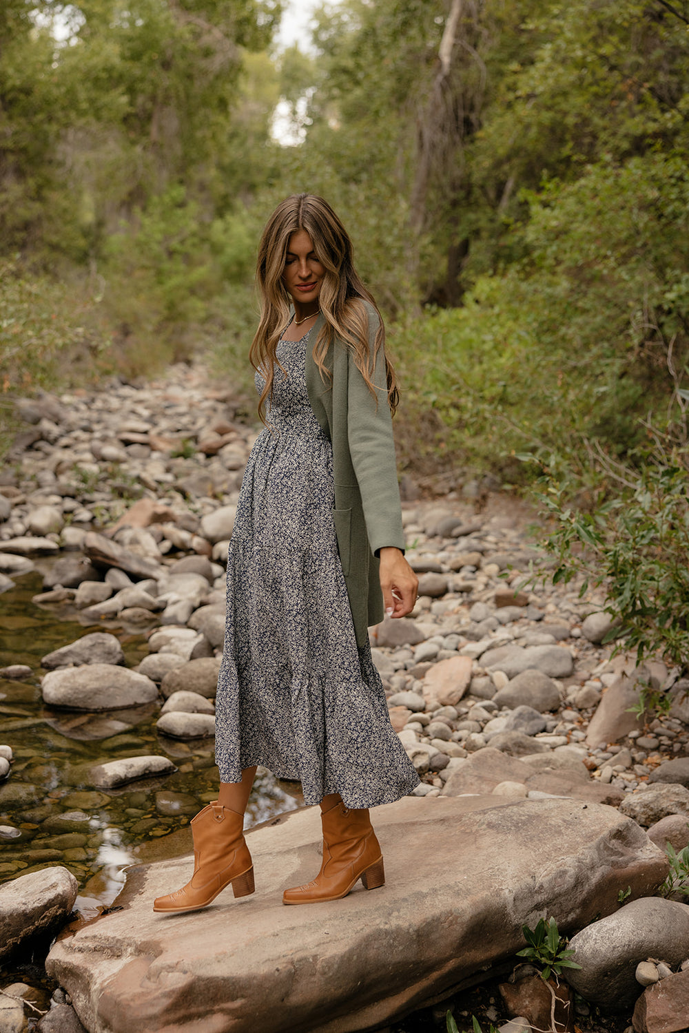 Woman in a floral dress and green cardigan standing on rocks by a stream with trees in the background