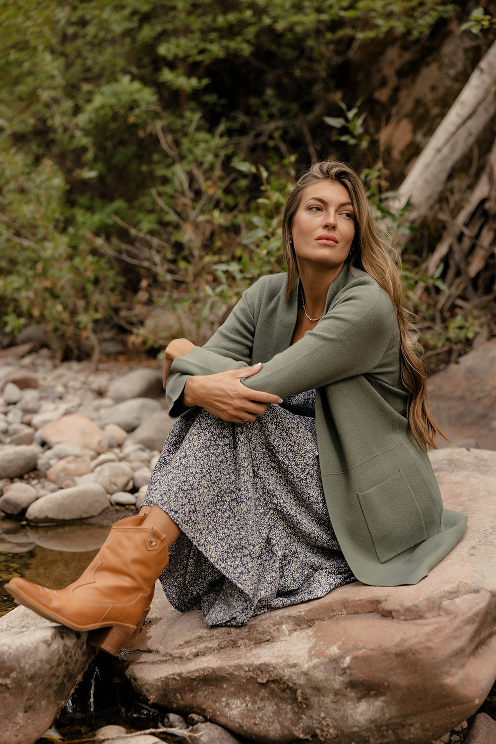 Woman sitting on a rock by a stream wearing a green coat and brown boots.