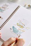 Person drawing a teapot illustration in a sketchbook with paint tubes nearby.