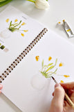Person drawing flowers in a notebook with a paintbrush, surrounded by art supplies on a white surface.