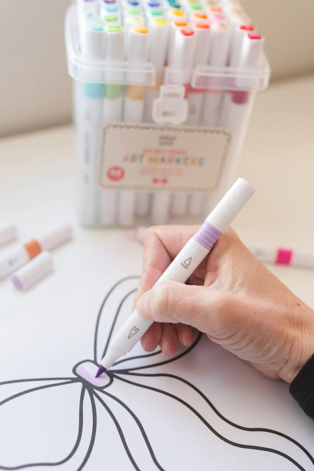 Hand holding a white marker with purple cap, coloring a butterfly on paper, with a container of markers in the background.