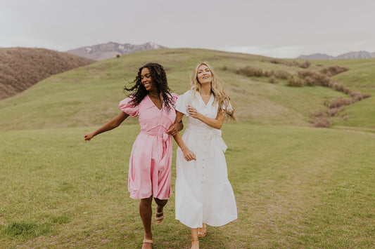 Two women walking along a hillside with linked arms. One is in a pink dress, and one is in a white dress.