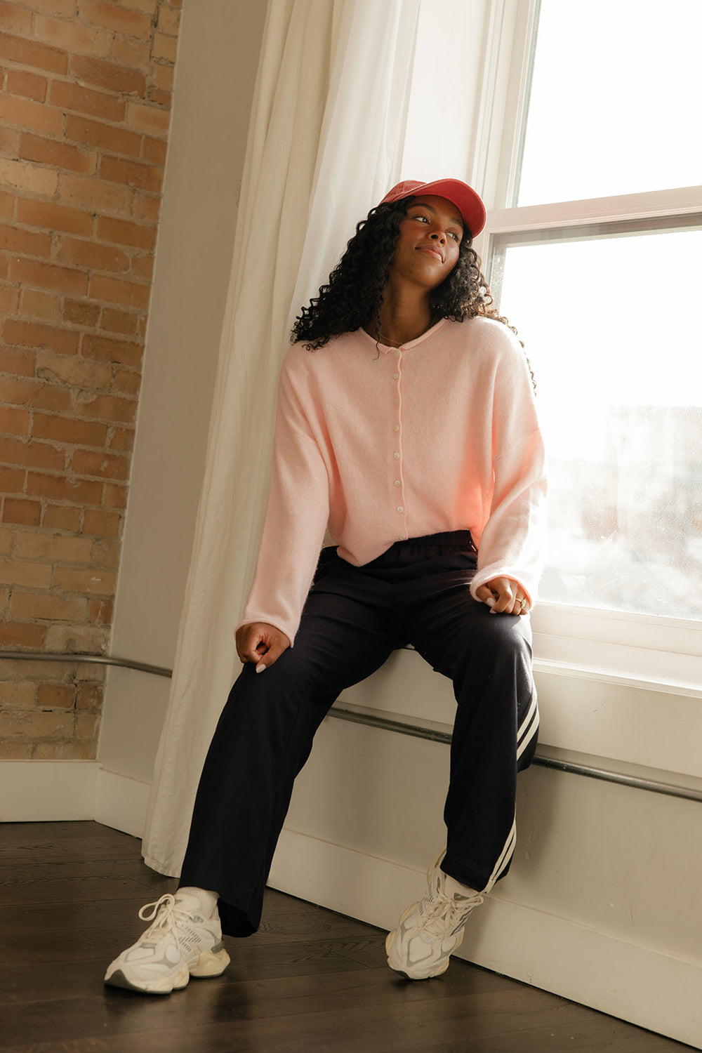 Woman sitting on a windowsill wearing a pink sweater and black pants.