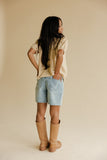 Woman wearing a beige shirt, light blue shorts, and tan boots against a beige background
