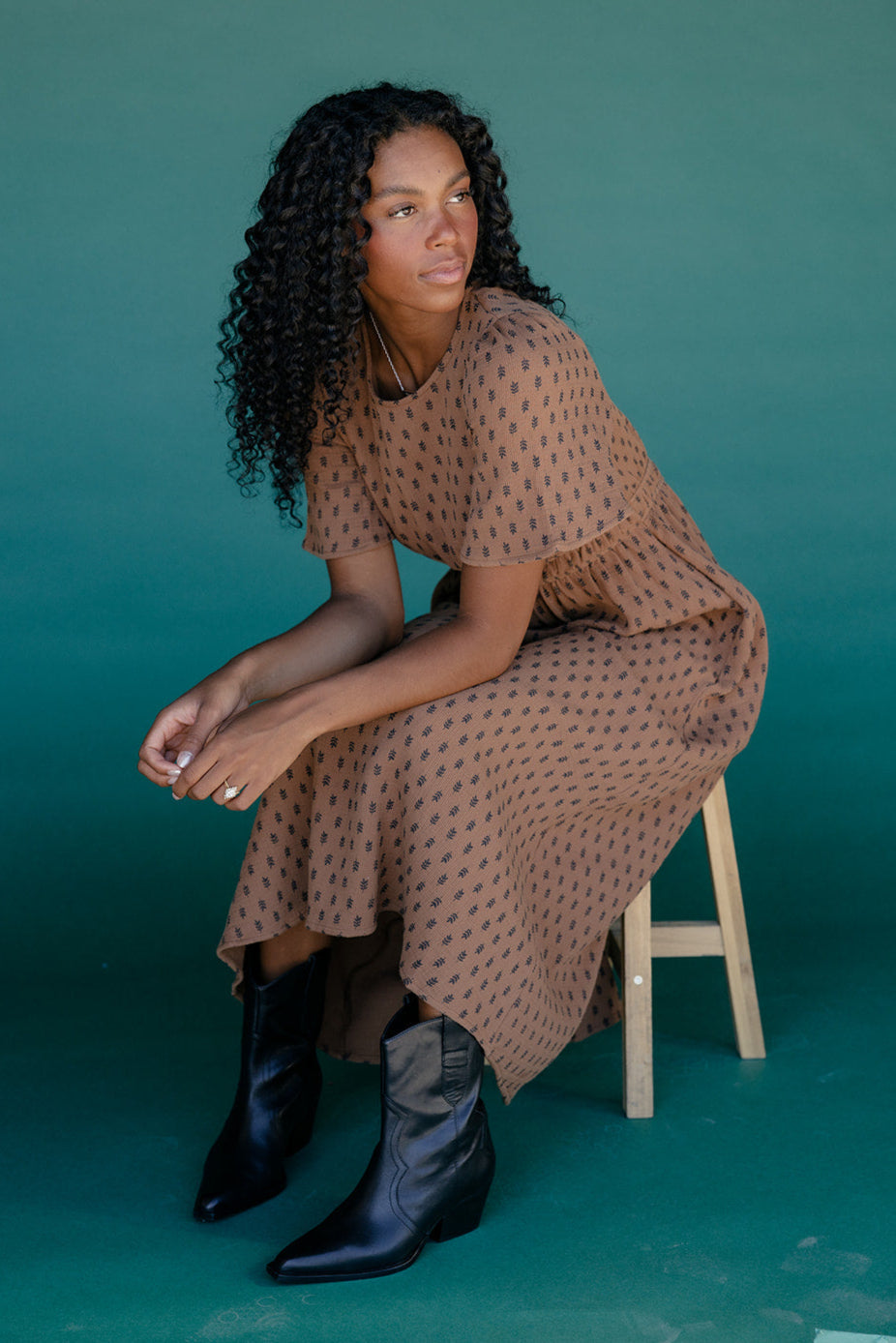 Woman sitting on a stool wearing a brown patterned dress and black boots against a teal background