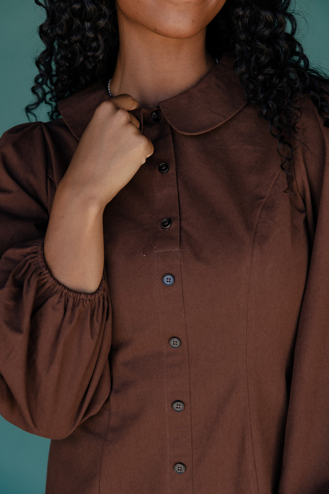 Brown shirt with long sleeves and buttons being adjusted against a teal background