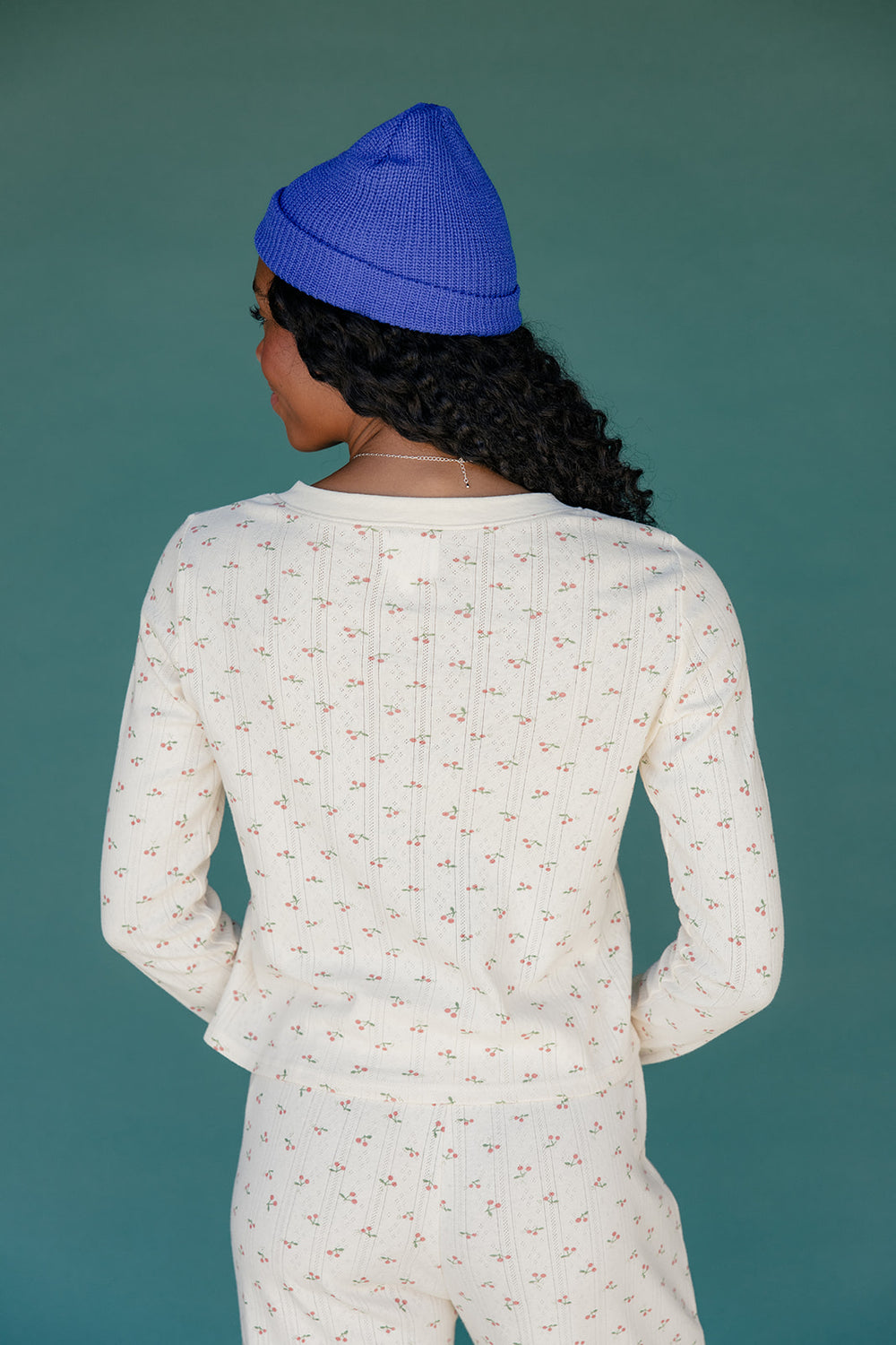 Person wearing a blue beanie and white patterned outfit against a teal background