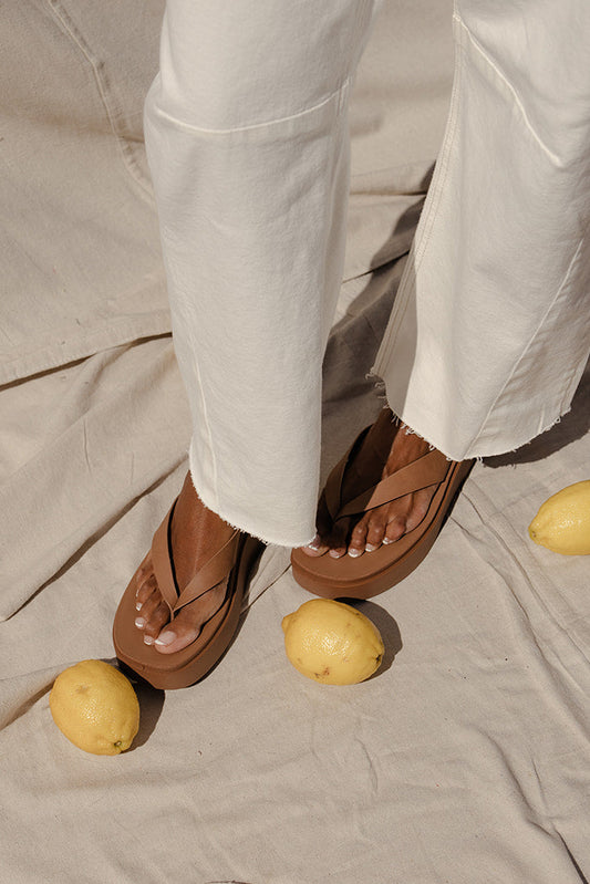 Person wearing white pants and brown sandals standing on a beige fabric surface with lemons scattered around.