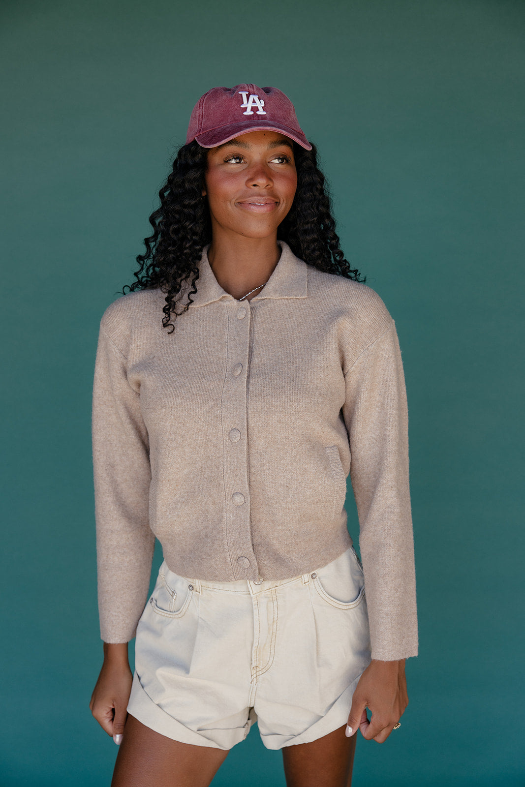 Woman wearing a beige button-up shirt and white shorts with a maroon cap against a teal background