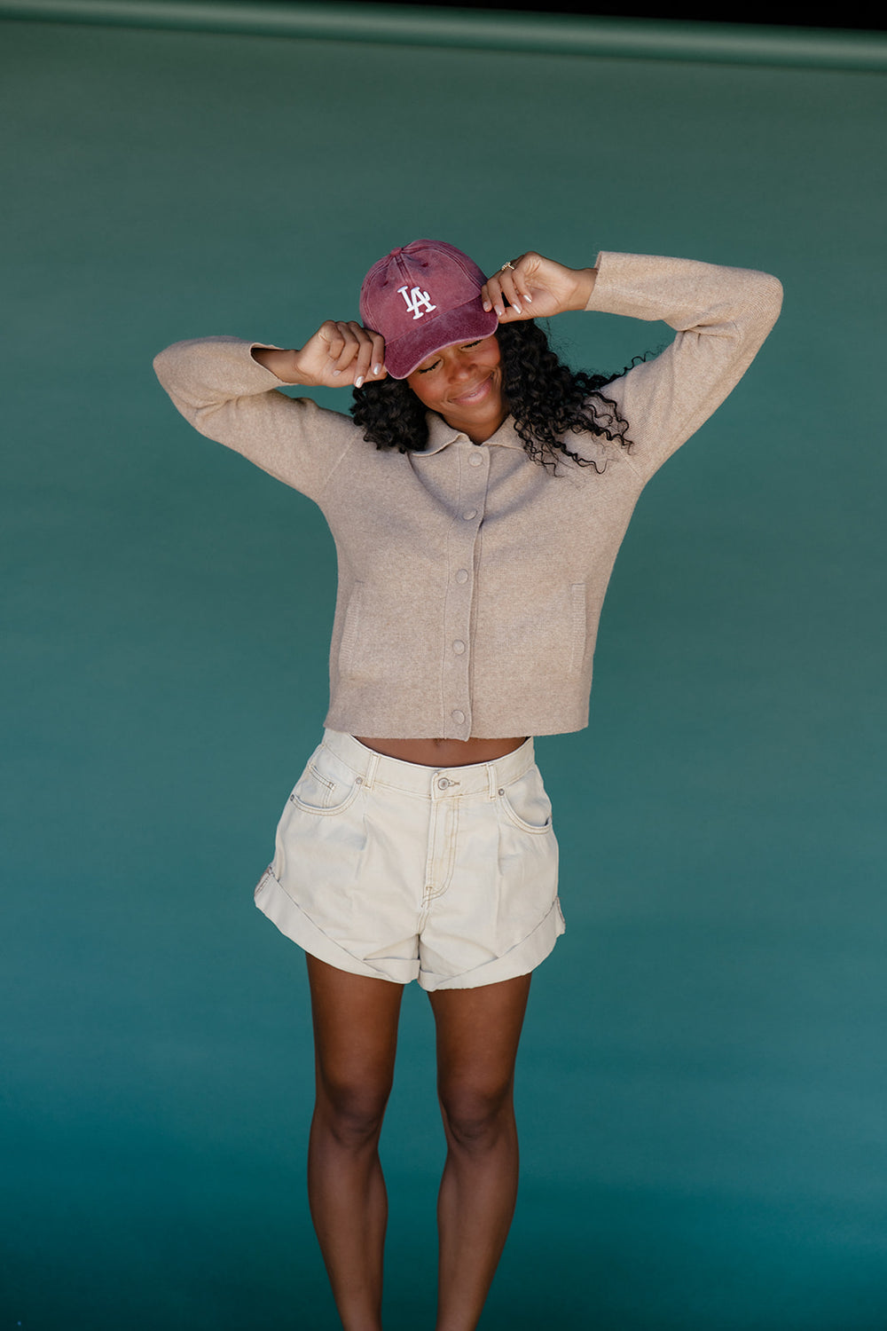 Person wearing a beige jacket and white shorts with a purple cap against a teal background