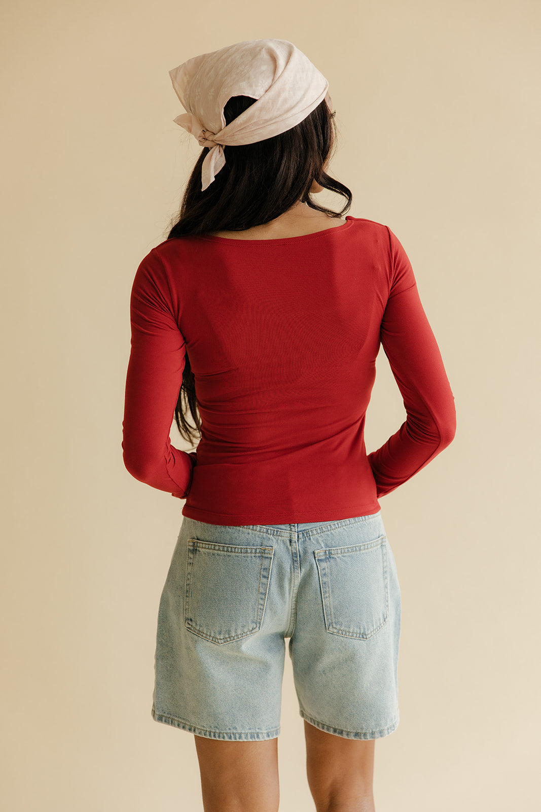 Person wearing a red long-sleeve top and denim shorts with a beige headscarf on a beige background
