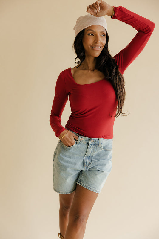 Woman wearing a red long-sleeve top and denim shorts against a beige background