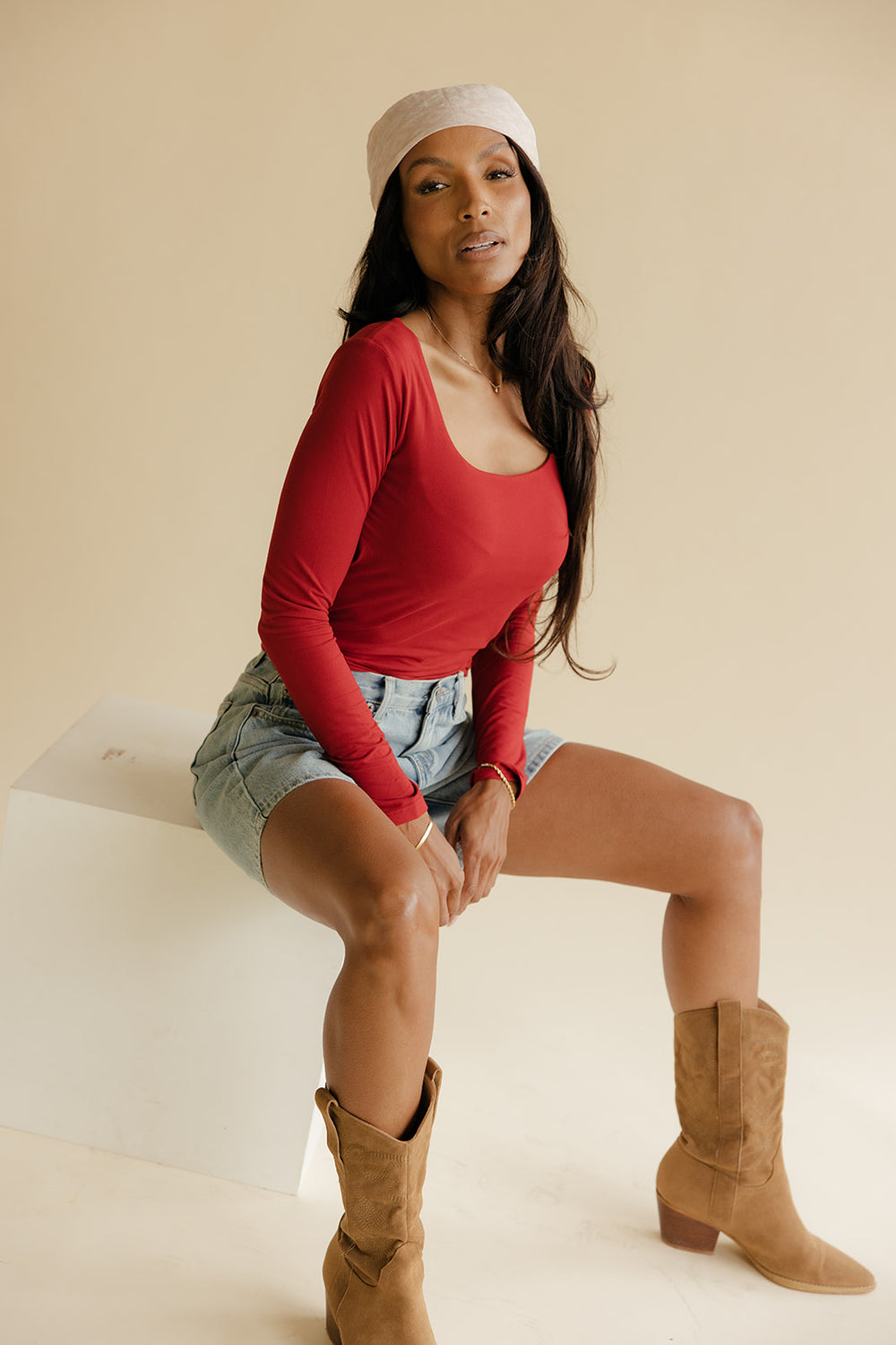 Woman wearing a red top, denim shorts, and tan boots sitting on a white surface.