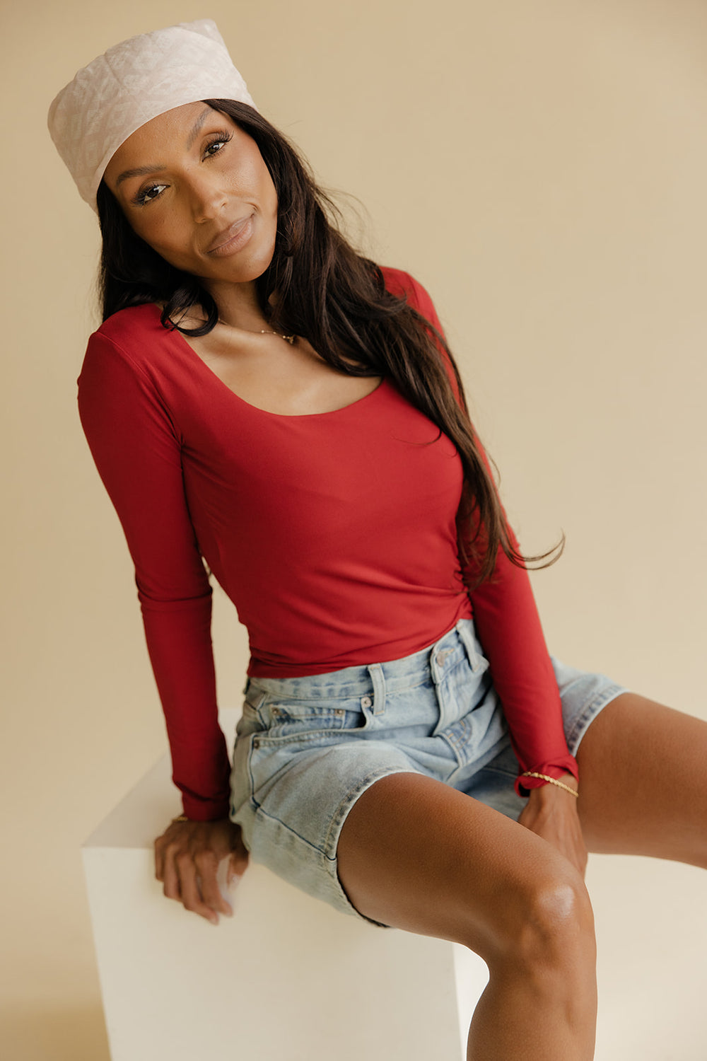Woman wearing a red long-sleeve top and denim shorts sitting on a white box against a beige background