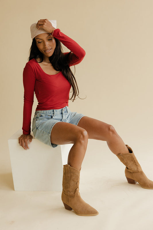 Woman wearing a red long-sleeve top, denim shorts, and brown boots sitting on a white stool against a beige background.