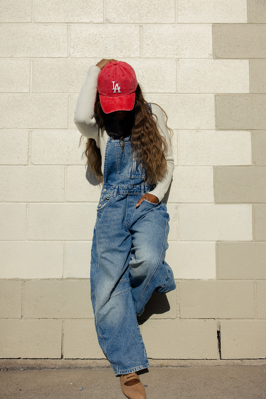 a girl in overalls and a red hat leaning against a wall