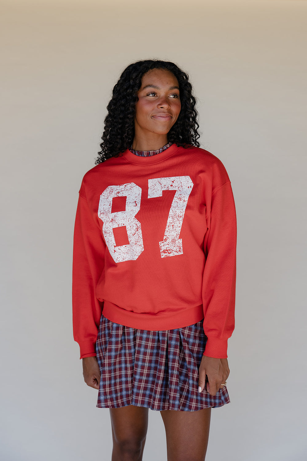 Person wearing a red sweatshirt with '87' and a plaid skirt against a plain background