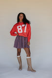 Person wearing a red sweatshirt with the number 87, plaid skirt, and brown boots on a plain background