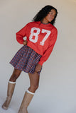 Woman wearing a red sweatshirt with '87', plaid skirt, and brown boots against a plain background