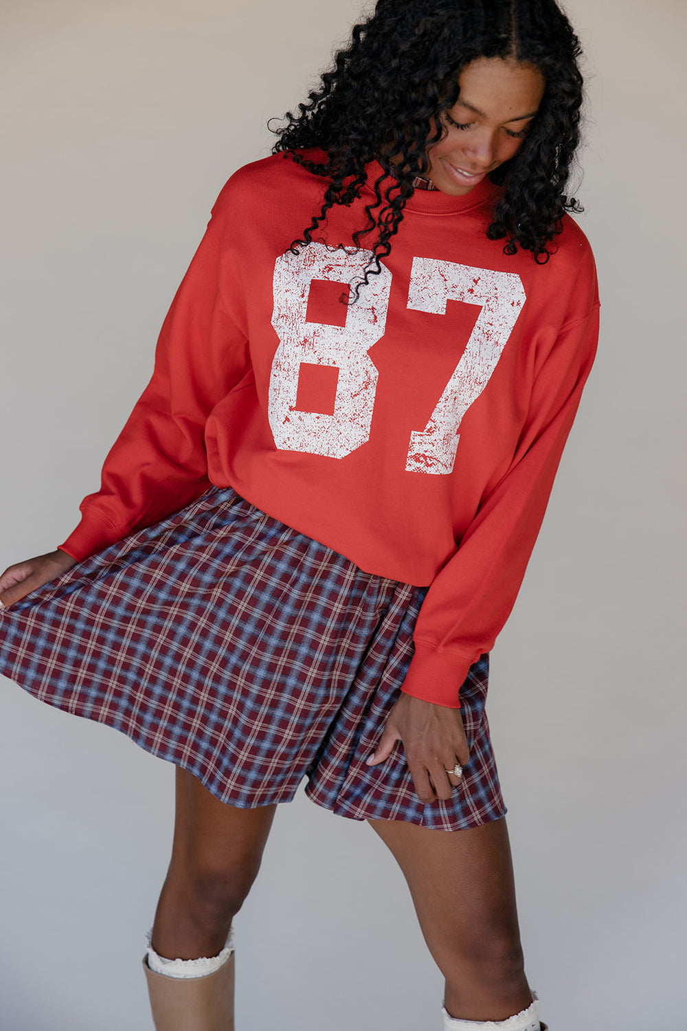 Person wearing a red sweatshirt with the number 87 and a plaid skirt against a plain background