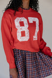 Person wearing a red sweatshirt with the number 87 printed on it, standing against a plain background.