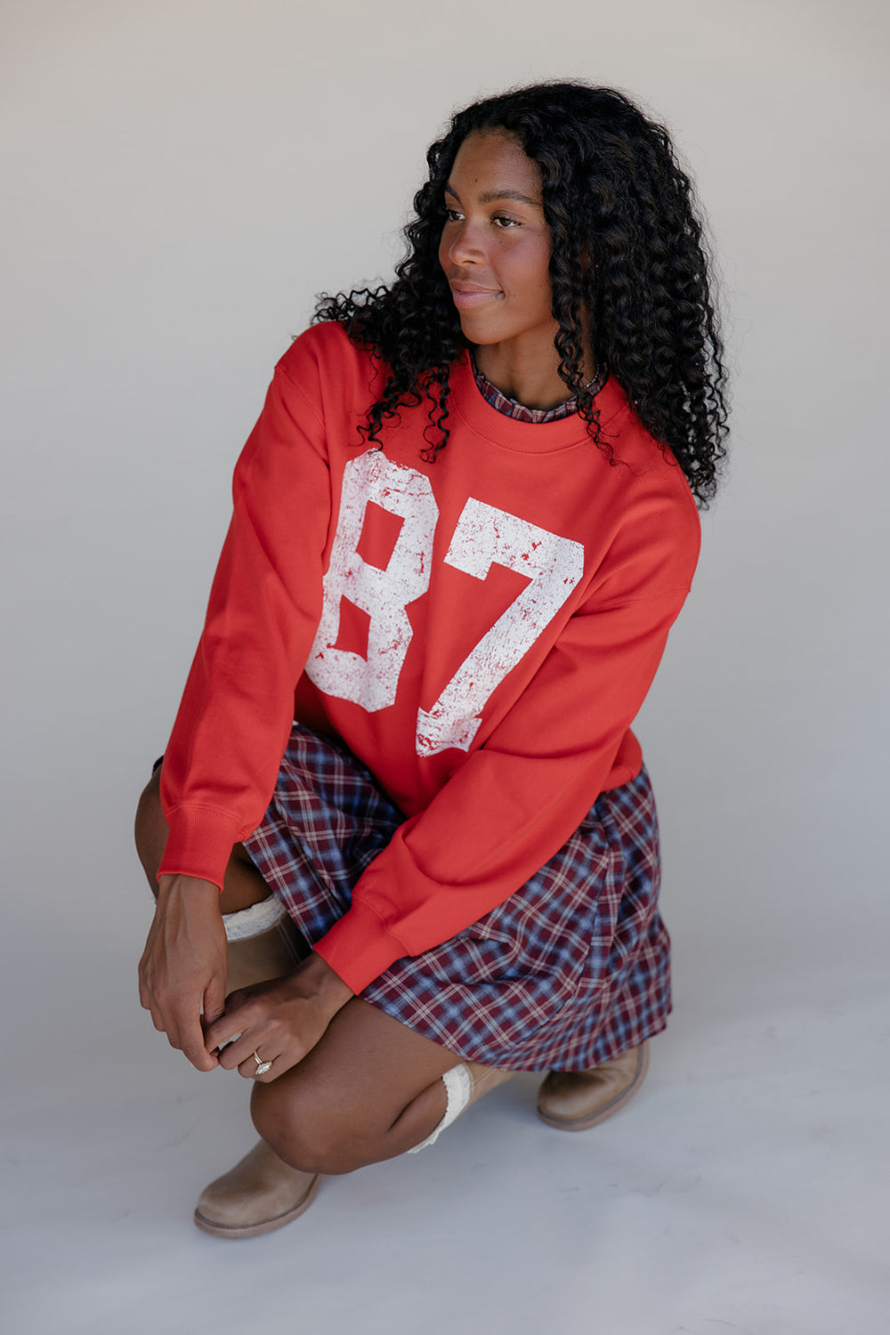 Woman wearing a red sweatshirt with the number 87 and a plaid skirt on a white background