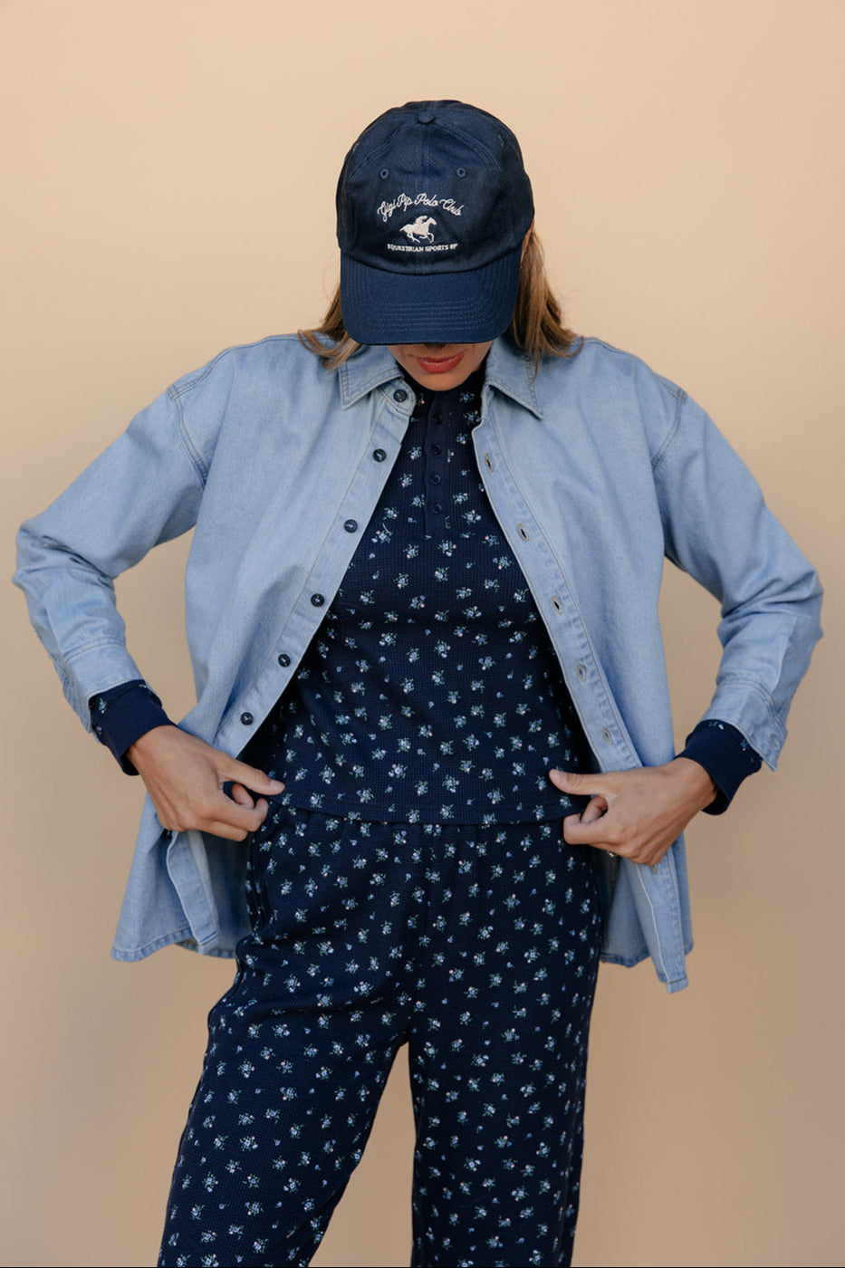 Person wearing a blue denim shirt over a navy outfit with a matching cap on a beige background