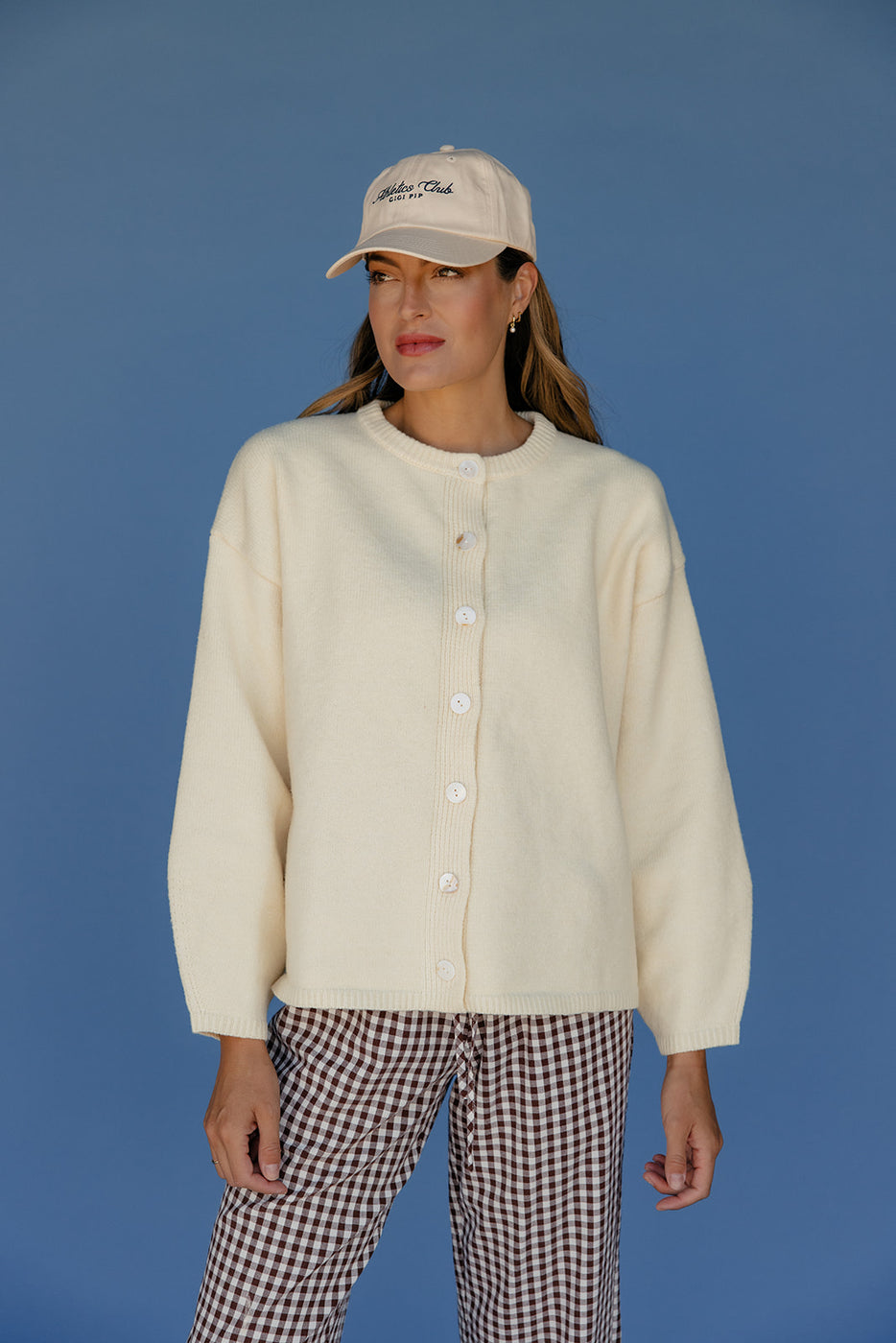 Person wearing a cream-colored cardigan, checkered pants, and a cap against a blue background