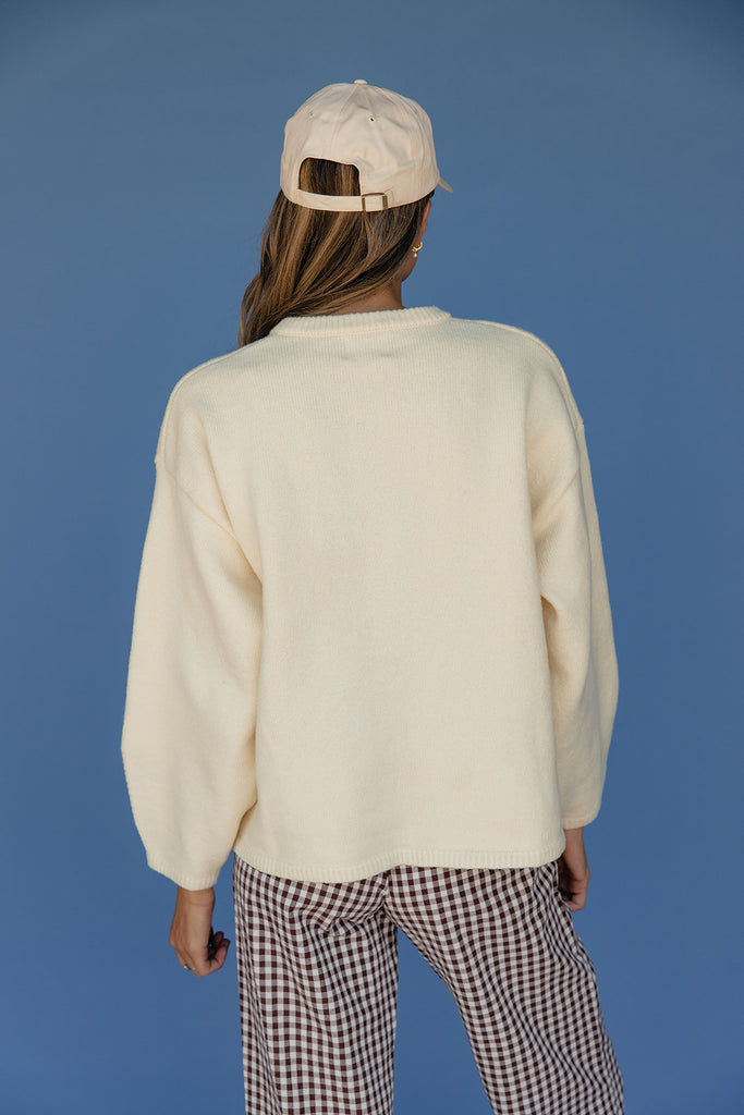 Person wearing a cream sweater and checkered pants with a beige cap on a blue background