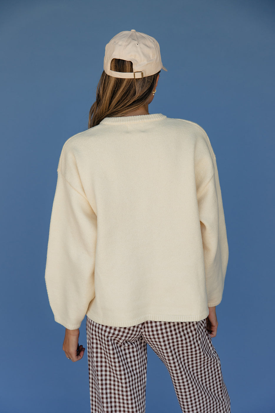 Person wearing a cream sweater and checkered pants with a beige cap on a blue background