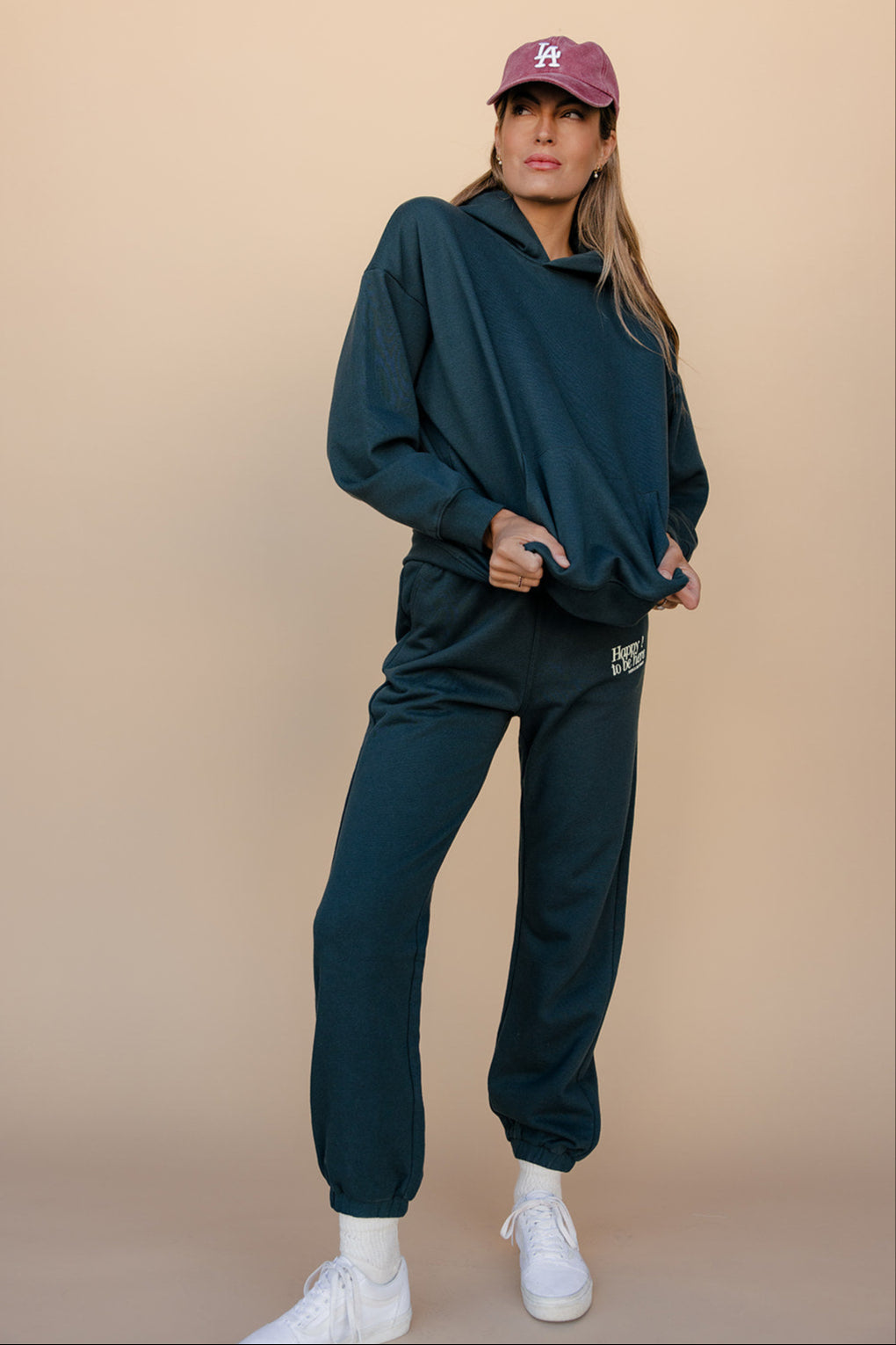 Person wearing a teal hoodie and pants with a pink cap on a beige background