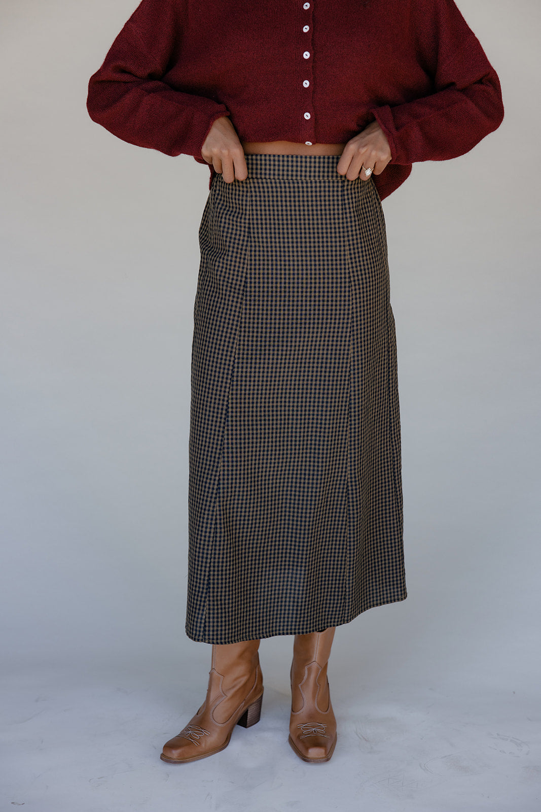 Person wearing a burgundy button-up shirt and plaid skirt with brown shoes on a plain background