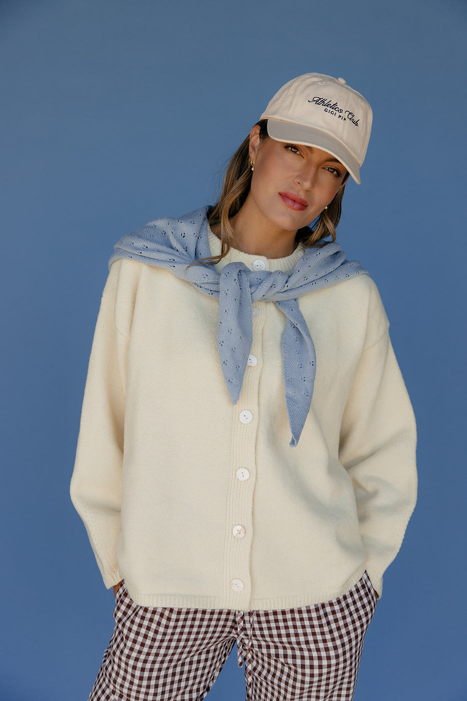 Person wearing a beige cardigan, blue scarf, and checkered pants against a blue background