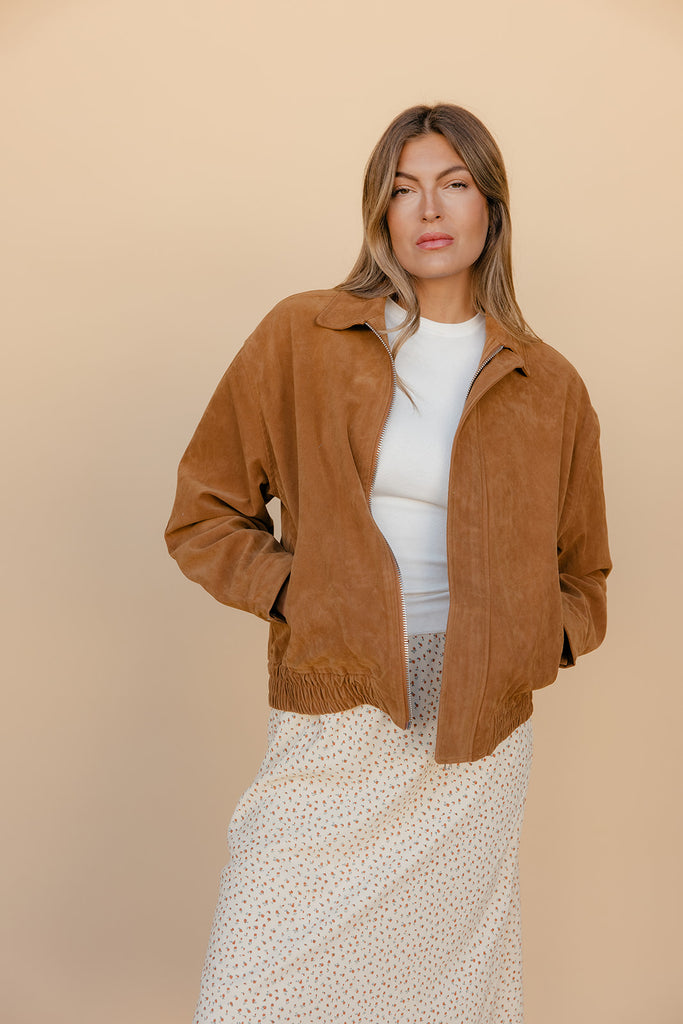 Woman wearing a brown jacket over a white top and patterned skirt on a beige background