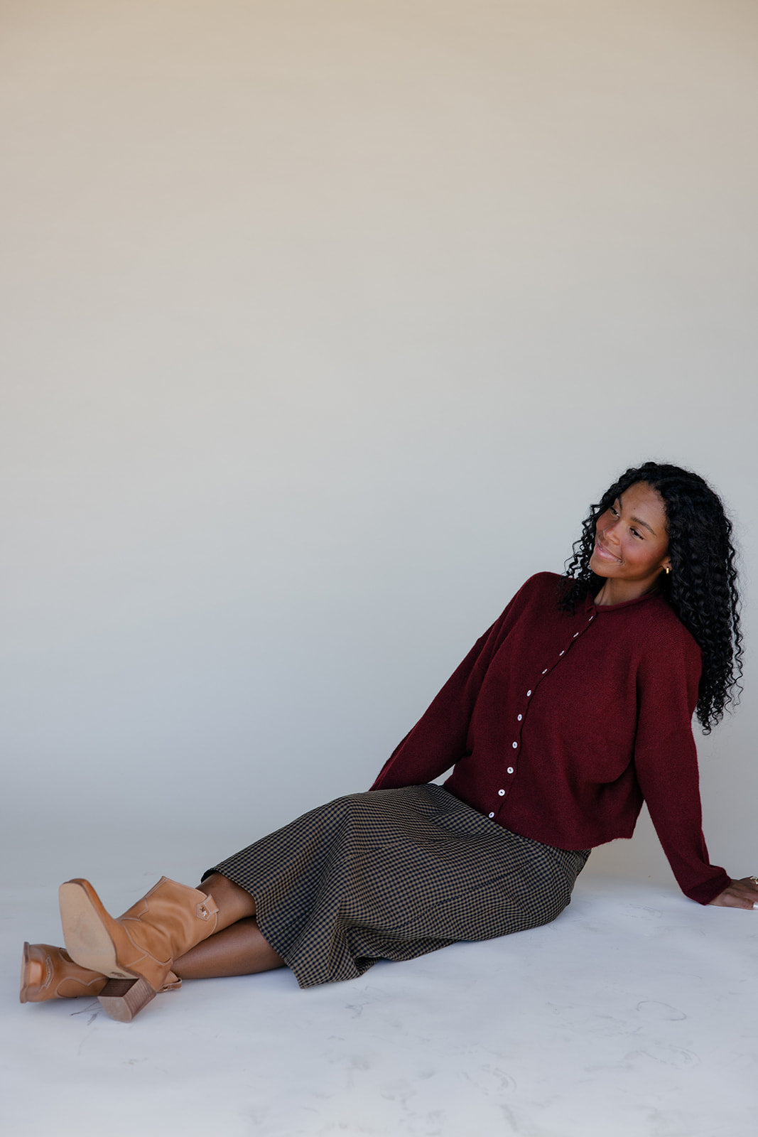 Woman sitting on a white surface wearing a burgundy sweater and patterned skirt.